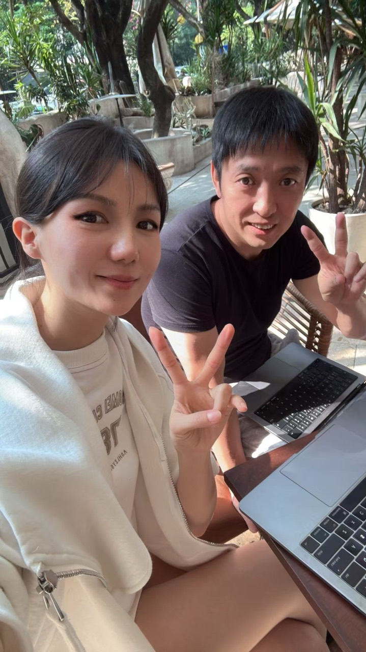 Miranda 和 Yan 在树荫下的户外咖啡店一起比耶自拍，桌上放着笔记本电脑